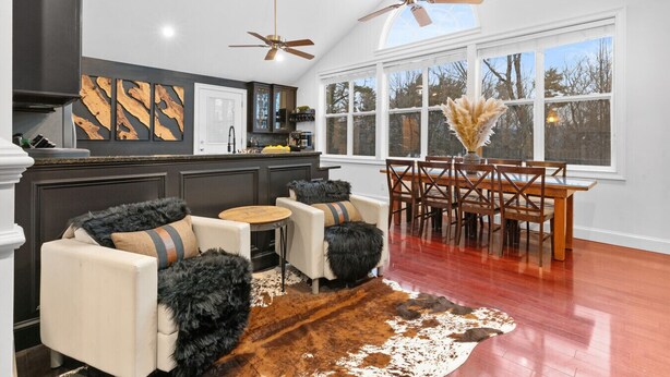 A modern kitchen with dark cabinetry, a dining area with a wooden table and chairs, and a large window with a view of trees. - Wild, Wild West Side! Hot tub, Games, Dog Friendly (Conway)