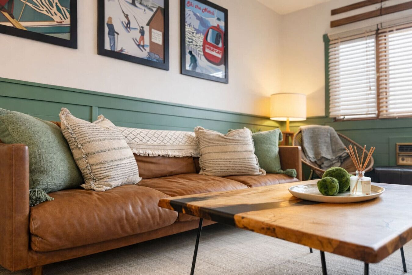 Una sala de estar acogedora con un sofá de cuero marrón, cojines verdes, una mesa de centro de madera y una lámpara.