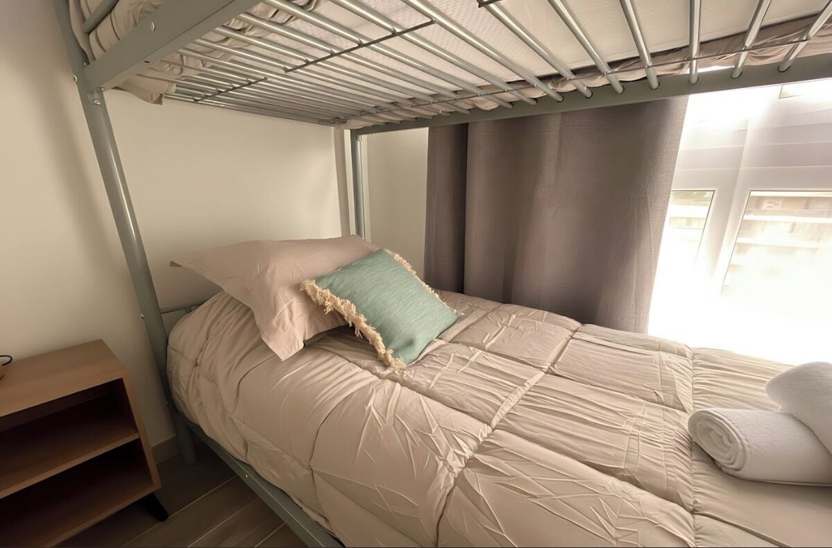 Una cama litera con colchón y almohadas color beige, una mesita de noche de madera y una ventana con cortinas.