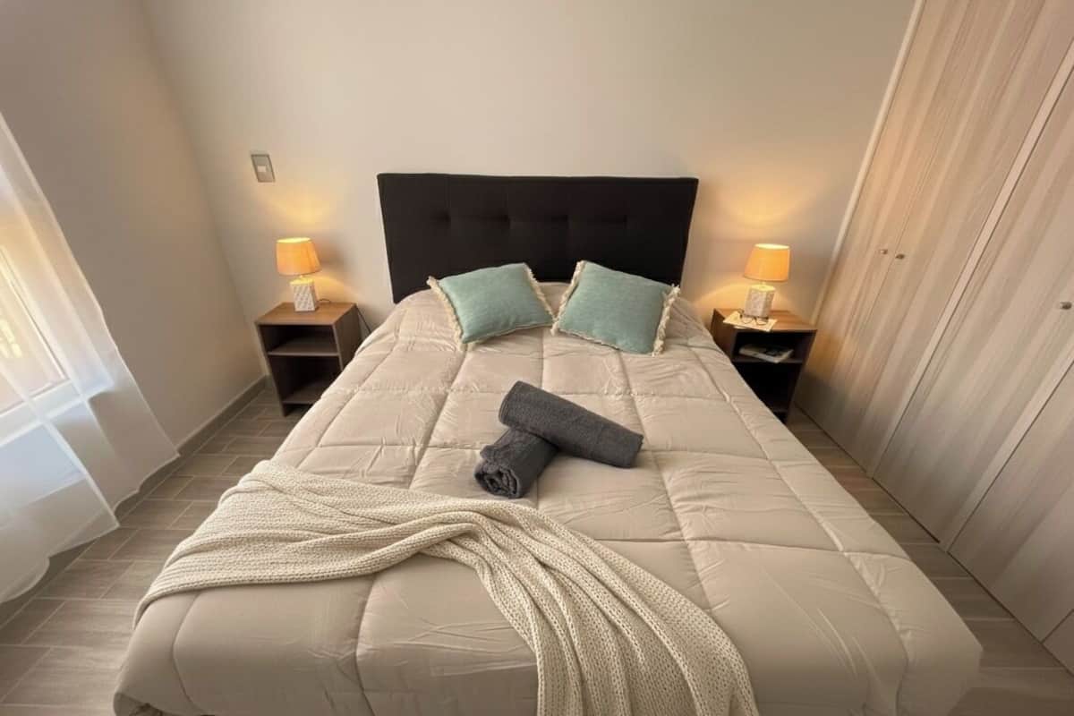 Een keurig opgemaakt bed met een gewatteerd hoofdbord, twee kussens en een opgerolde handdoek. Twee nachtkastjes met lampen, en een raam met dunne gordijnen aan de linkerkant.