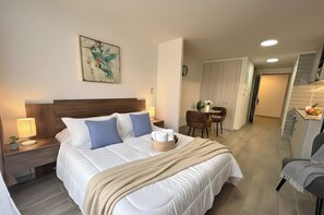 A hotel room with a large bed, a wooden headboard, a bedside table with a lamp, a small potted plant, and a painting on the wall. - Movistar Arena Santiago - Domus Travels (Santiago)