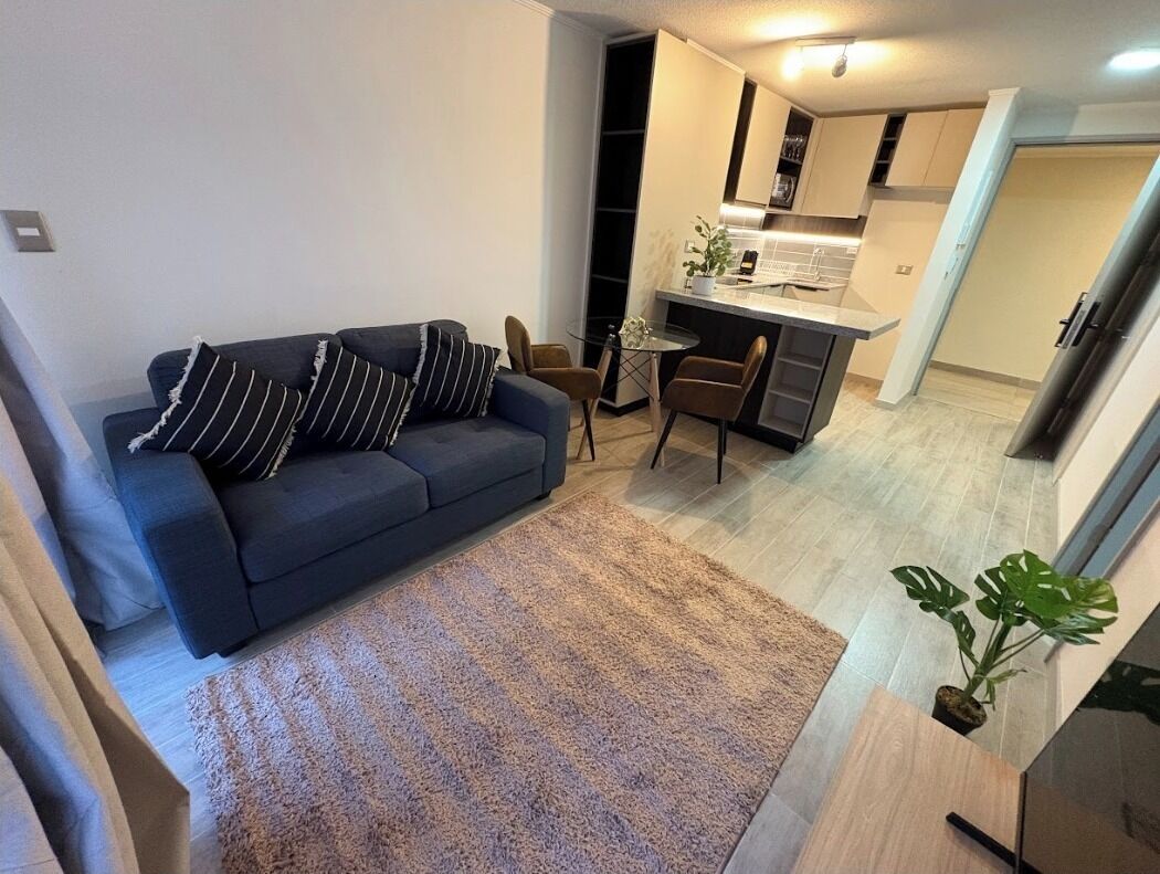 Una sala de estar moderna con un sofá azul oscuro, almohadones a rayas y un mullido tapete marrón. Una pequeña mesa de vidrio con sillas se encuentra en el centro. Se ve una cocina al fondo, con armarios blancos y encimera negra.