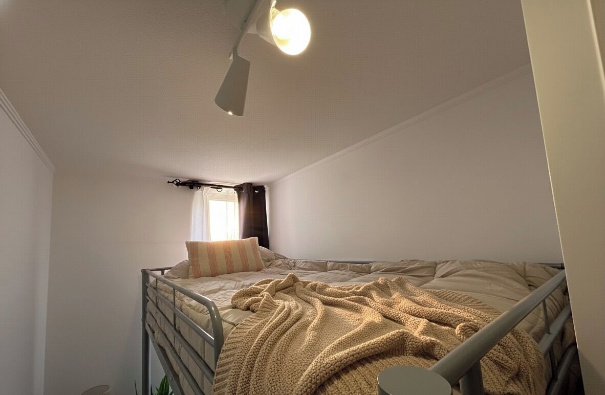 Ett litet rum med en enkel säng, en våningssäng, en takfläkt och ett fönster med gardiner.
