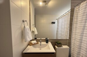 A modern bathroom with a large mirror, white tiles, a sink with a soap dispenser, and a shower curtain. - Movistar Arena Santiago - Domus Travels (Santiago)