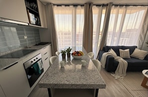 Une cuisine moderne dotée d’un plan de travail en granit, d’un four et d’un coin repas avec une table dressée pour deux personnes. Le salon comprend un canapé de couleur foncée et offre une vue sur les bâtiments grâce à de grandes fenêtres.