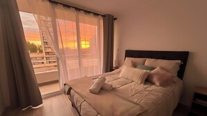 A bedroom with a large bed, a view of the sunset through sheer curtains, and a balcony with a railing. - Movistar Arena Santiago - Domus Travels (Santiago)