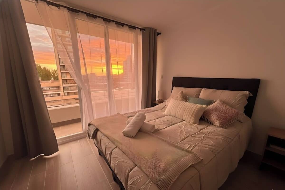 Een slaapkamer met een groot bed, uitzicht op de zonsondergang door dunne gordijnen en een balkon met een hekwerk.