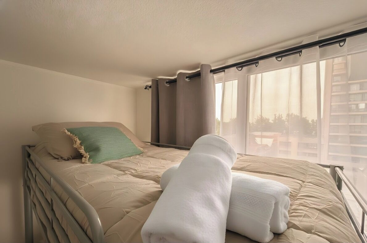 A single bed with a beige mattress, a green pillow, and rolled white towels.
