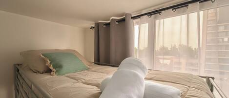 A single bed with a beige mattress, a green pillow, and rolled white towels.
