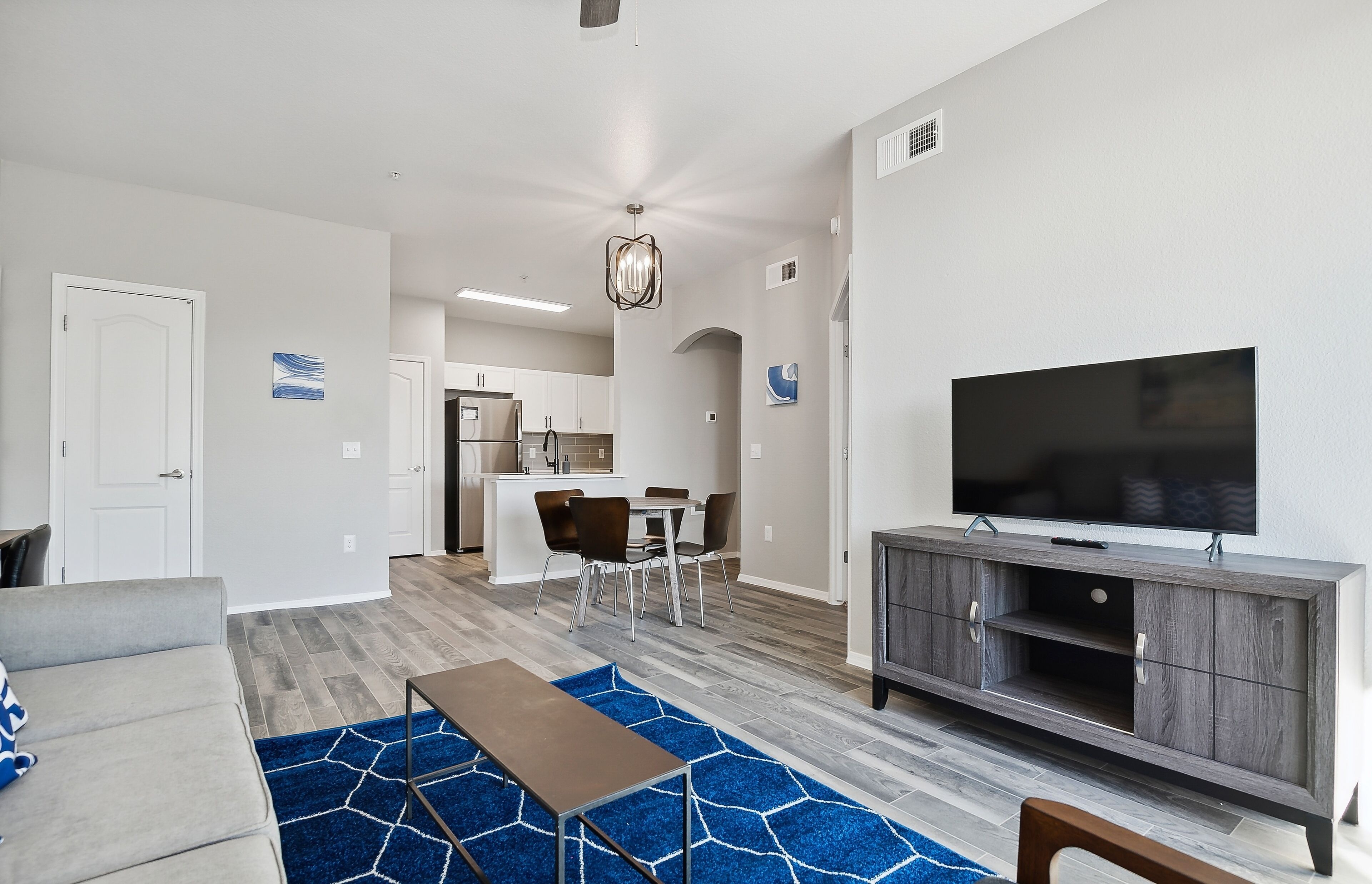 A modern living room with a flat-screen TV, grey sofa, and a dining area with chairs and a table.