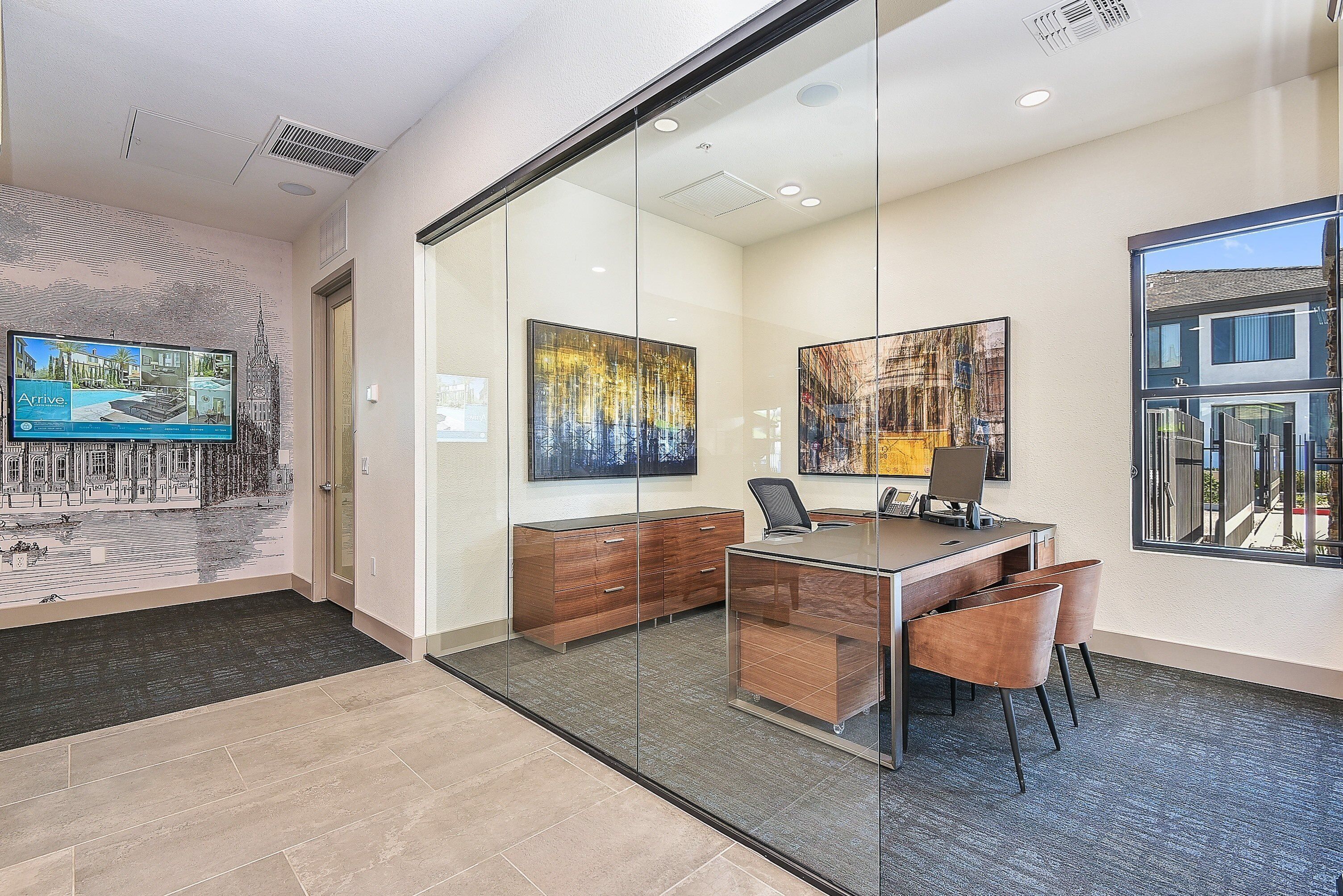 A modern office space with a glass partition, a wooden desk, two chairs, and artwork on the walls.