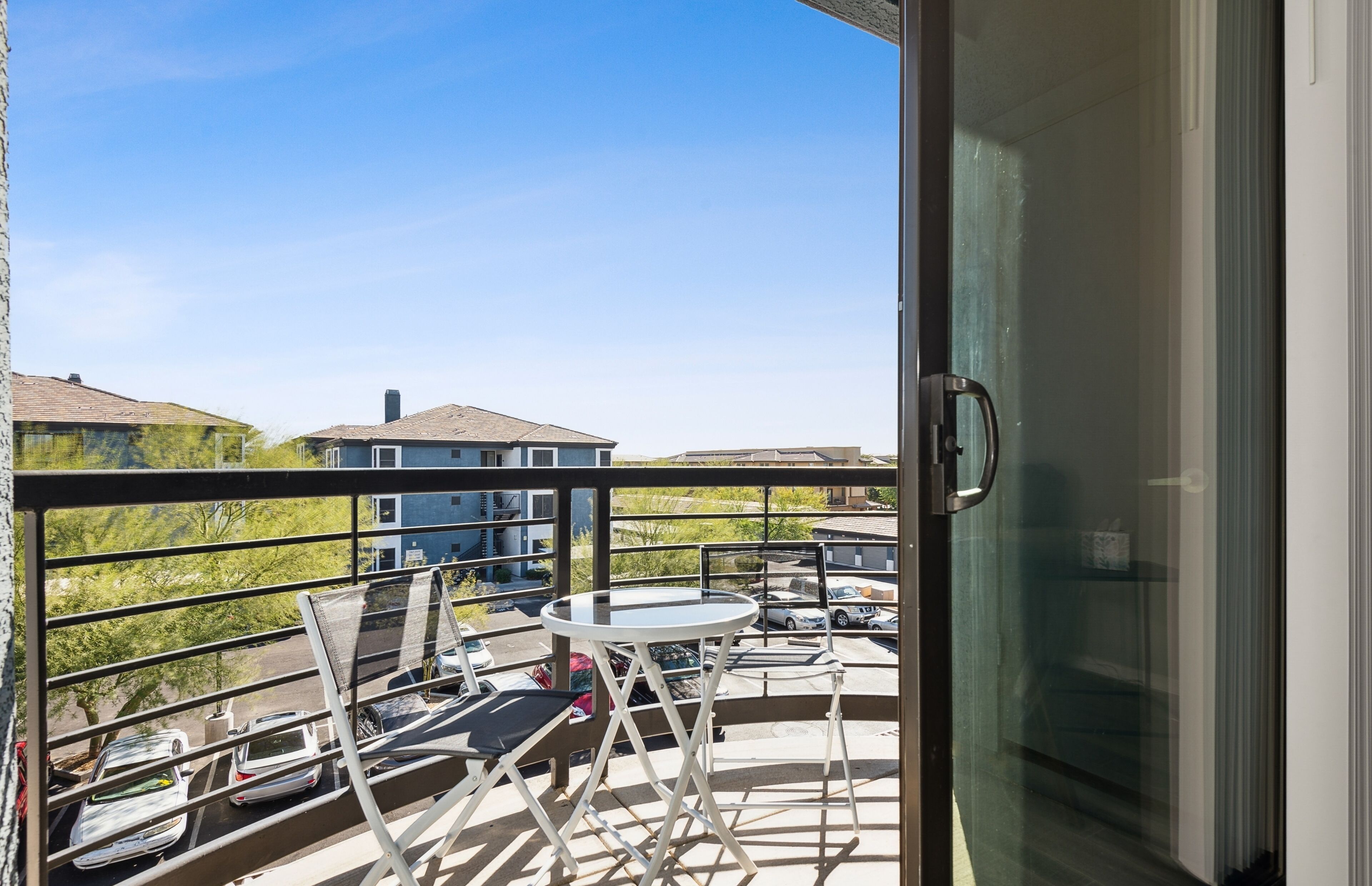A balcony with a table and chairs, a glass door, and a view of parked cars and buildings.