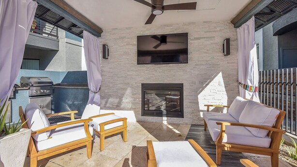 Un patio al aire libre con un ventilador de techo, un televisor de pantalla plana montado en una pared de piedra, una chimenea y sillones de madera.