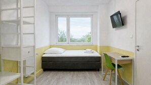 A small, well-lit room with a bed, a desk with a chair, a TV mounted on the wall, and a window providing a view of the outdoors. - Silvio-Gesell-Tagungsstätte (Wuppertal)