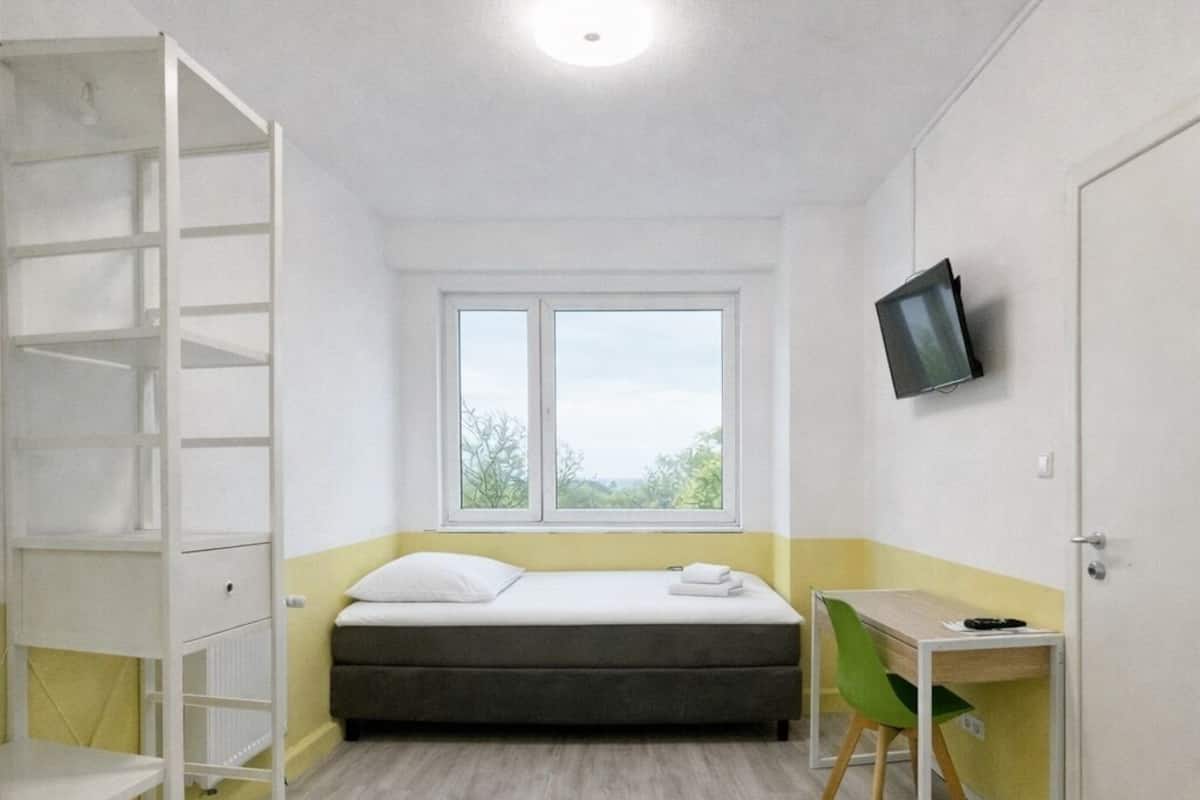 A small, well-lit room with a bed, a desk with a chair, a TV mounted on the wall, and a window providing a view of the outdoors.