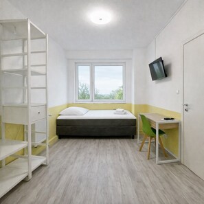 Una stanza piccola e ben illuminata con un letto, una scrivania con una sedia, una TV montata a parete e una finestra che offre vista sull'esterno.
