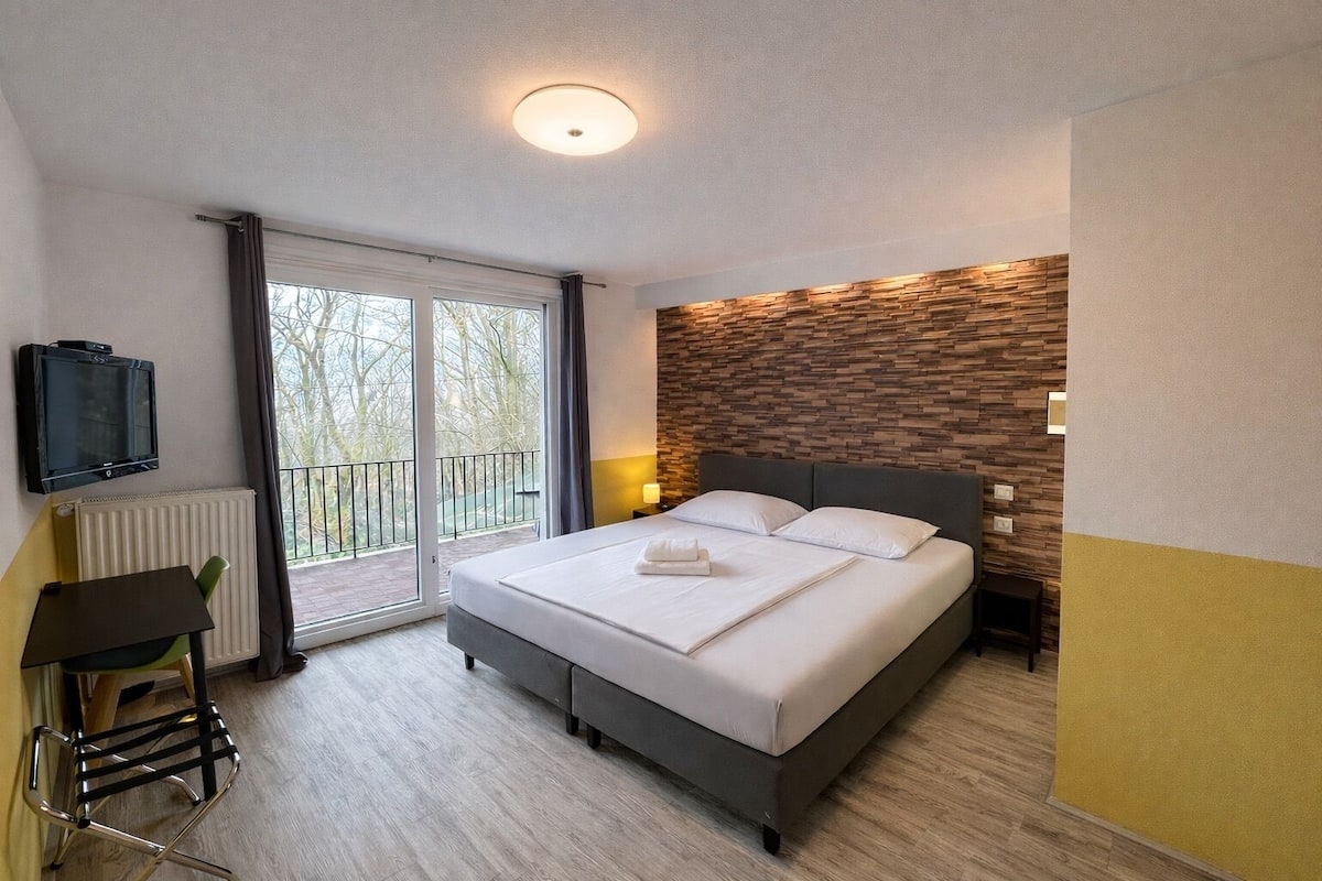 A modern hotel room with a large bed, a TV, a desk, and a balcony with a view of trees.