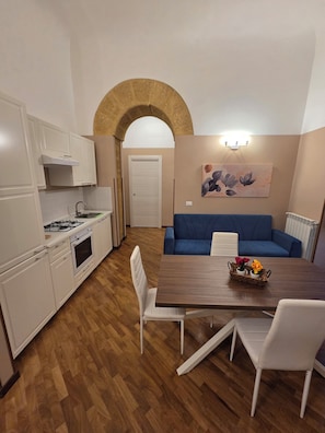 A compact kitchen with white cabinets, a stove, and a sink. There is a dining area with a wooden table and blue sofa. - Family apartment -Civico39Home (Agrigento)