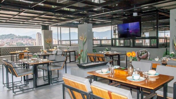 En moderne restaurant med høje lofter, store vinduer og udsigt over byen. Bordene er dækket med tallerkener, glas og bestik.