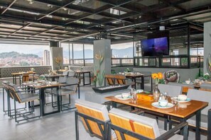A modern restaurant with high ceilings, large windows, and a city view. Tables are set with plates, glasses, and cutlery. - Furnished 2-room apartment Apartment at Edificio Helena in Medellín + Breakfast (Medellín)