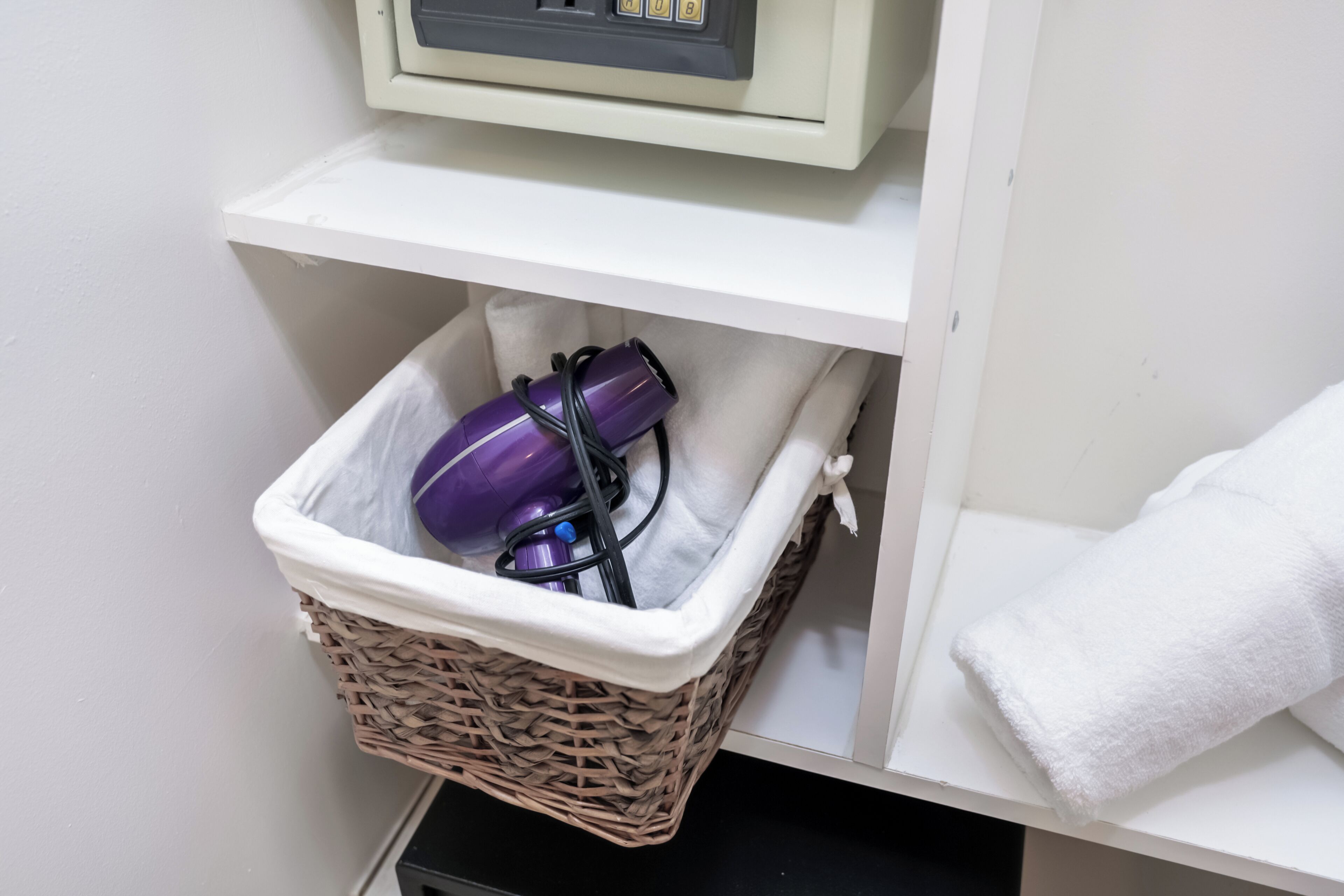 A bathroom with a white shelf, a wicker basket containing a purple hairdryer and its cord, and a white towel hanging on the wall.