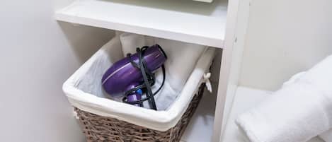 A bathroom with a white shelf, a wicker basket containing a purple hairdryer and its cord, and a white towel hanging on the wall.