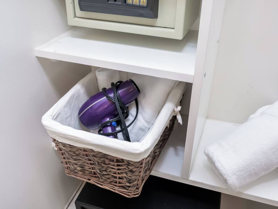 Une salle de bain avec une étagère blanche, un panier en osier contenant un sèche-cheveux violet et son cordon, et une serviette blanche accrochée au mur.