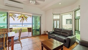 Ein Wohnzimmer mit einer Ledersofa, einem Holztisch, Korbstühlen und einer Glastür, die auf einen Balkon mit Blick auf Strand und Meer führt.