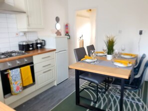 A modern kitchen with white cabinets, a stove, and a small refrigerator. There is a dining table set with plates, glasses, and a plant. - Spacious 3BR - Nr Clifton Train Station 510min Walk - Garden & Free Parking (Manchester)