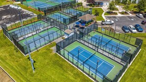 Luchtbeeld van een sportcomplex met meerdere tennisbanen, omgeven door groen en een parkeerplaats.