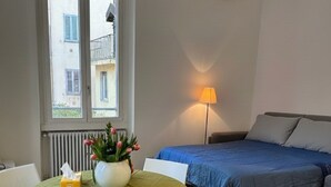 Een kleine, nette kamer met een bed, een tafel met stoelen, een lamp en een raam met uitzicht op gebouwen.