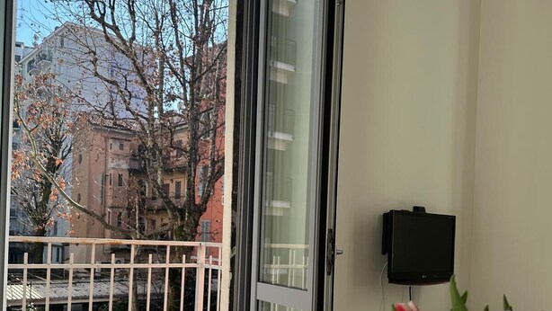 Een balkon met uitzicht op gebouwen en bomen, een witte tafel met een bloevas en een zwart object aan de muur.