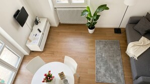 Een moderne woonkamer met een witte bank, een grijs vloerkleed, een eettafel met stoelen, een potplant en een tv op een dressoir.