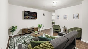 A modern living room with a sofa, armchairs, a coffee table, and a TV. There is a clock on the wall and a plant on the side.
