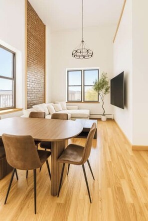 Een moderne woonkamer met een ronde houten tafel, comfortabele zithoek, een groot raam, een accentmuur van baksteen en een flatscreen televisie aan de muur bevestigd.