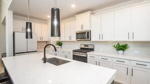 Cocina moderna con armarios blancos, una isla central con lavamanos, electrodomésticos de acero inoxidable e iluminación colgante.