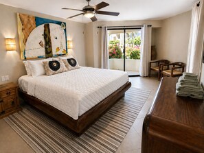 A bedroom with a bed, a ceiling fan, a wooden nightstand, a desk with towels, and a view of a balcony with plants. - Spacious 2BR 2BA Beachfront Walk-Out Condo in Terrasol Resort, Cabo San Lucas (Cabo San Lucas)