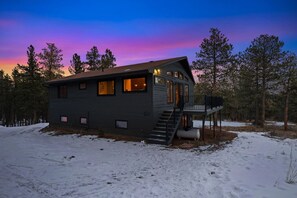 A two-story cabin with a deck, surrounded by trees and snow. - The Pine Getaway on 60 Acres with Hot Tub (Pine)