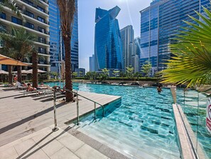 A swimming pool with clear water, surrounded by palm trees and lounge chairs, with tall buildings in the background. - Stayshort - 1 Bed in Business Bay With Modern Amenities (Dubai)