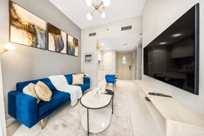 A modern living room with a blue sofa, a white coffee table, and a large flat-screen TV. - Stayshort - 1 Bed in Business Bay With Modern Amenities (Dubai)