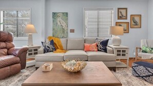 Ein Wohnzimmer mit einem beigefarbenen Ecksofa, einem Holztisch, und einem braunen Ledersessel. Zwei Nachttische mit Lampen, ein Bild an der Wand und ein Fenster mit Jalousien sind ebenfalls vorhanden.