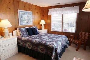 Ein Schlafzimmer mit einer holzgetäfelten Wand, einem Bett mit gemustertem Überwurf, zwei Nachttischlampen, einem Sessel und einem Fenster mit Jalousien.