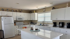 Una cocina moderna con armarios blancos, una isla central, un refrigerador, un microondas, un lavamanos y una ventana con persianas.