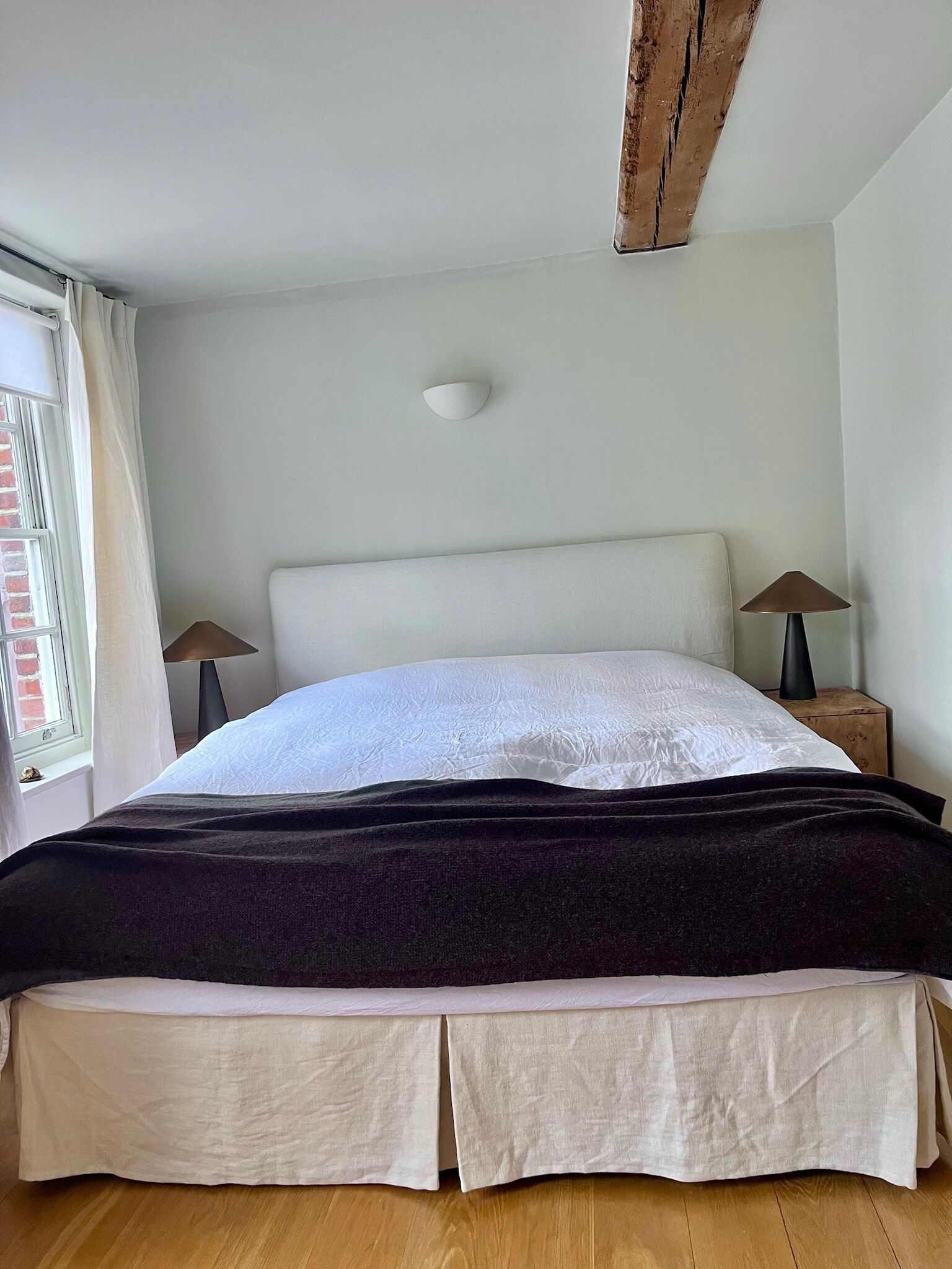 Un dormitorio con una cama grande, dos mesitas de noche con lámparas, un ventanal con cortinas y una viga de madera en el techo.