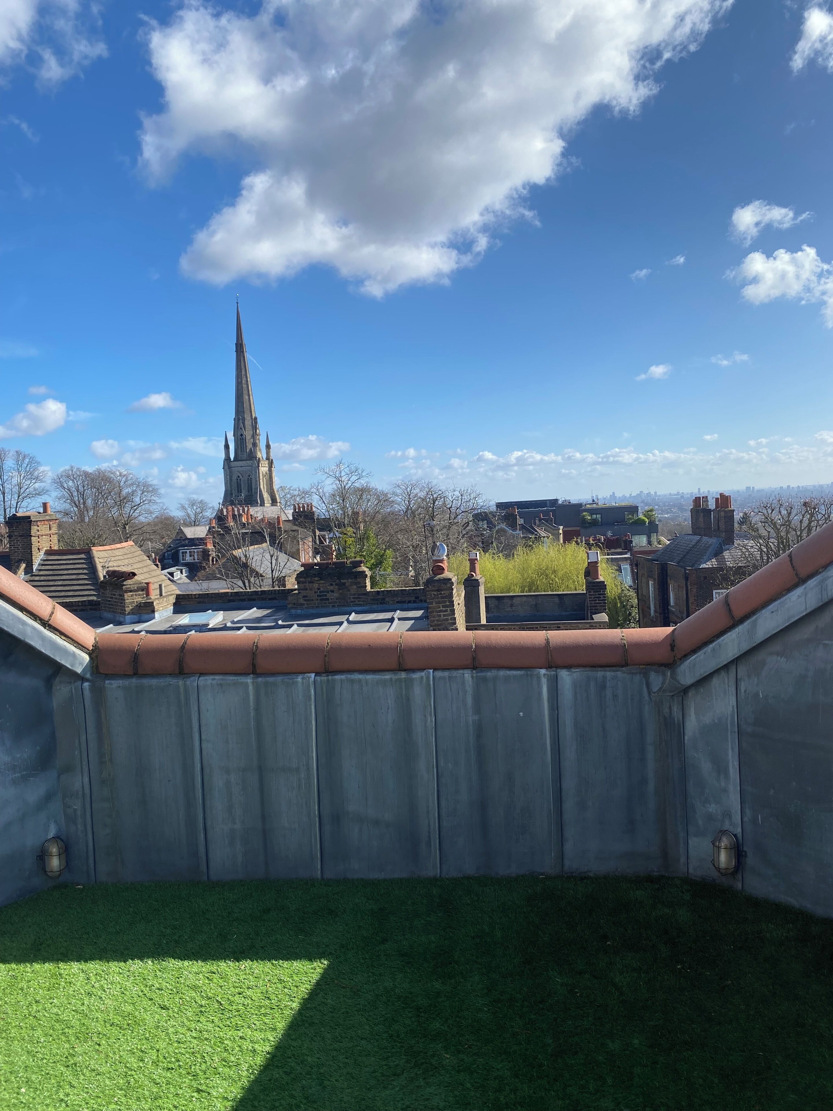 A rooftop terrace with a view of a church spire, a brick chimney, and a metal railing.