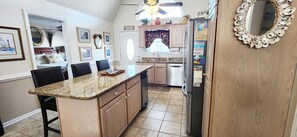 A kitchen with a granite countertop, wooden cabinets, a stainless steel refrigerator, and a dining area with chairs. - 5 Bedrooms Oceanfront Beach House for 10 (Ocean Isle Beach)