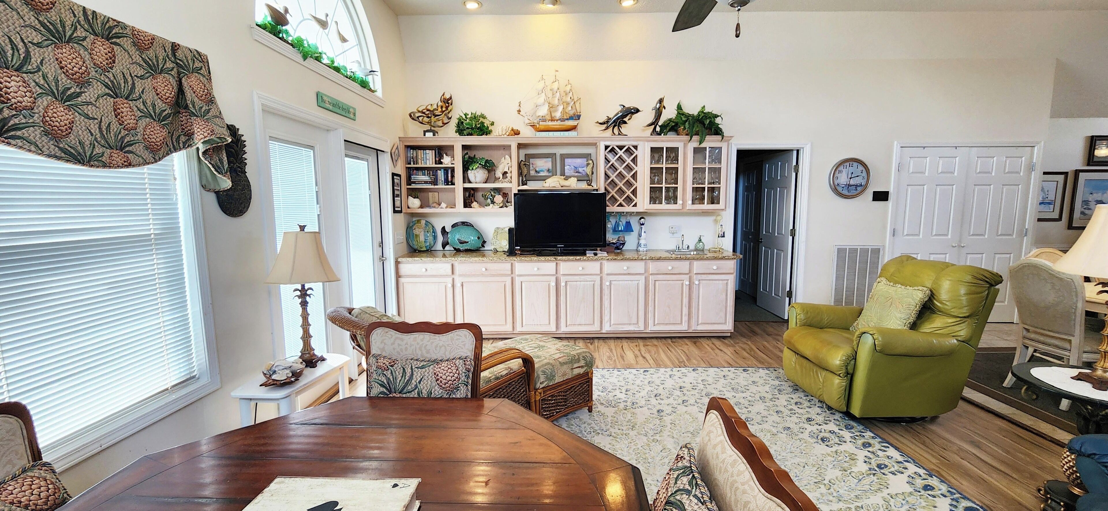 A dining area with a wooden table and chairs, a kitchen with a TV and decorative items, and a green armchair.