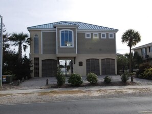 A two-story building with a gabled roof, a central entrance with a glass door, and two garage doors on the ground floor. - Turtle Time 4 Bedroom Community Pool (Ocean Isle Beach)