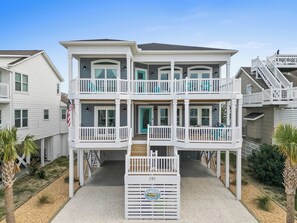 A two-story beachfront house with a balcony, a flag, and a number plaque. - New Luxury Beach House 5 Bedrooms Pool & Tiki Bar (Ocean Isle Beach)