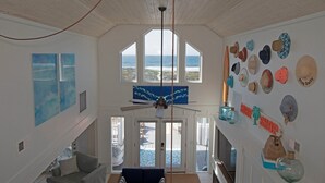 Una stanza con ventilatore a soffitto, ampie finestre con vista sull'oceano, una serie di cappelli appesi al muro e vista sulla spiaggia.